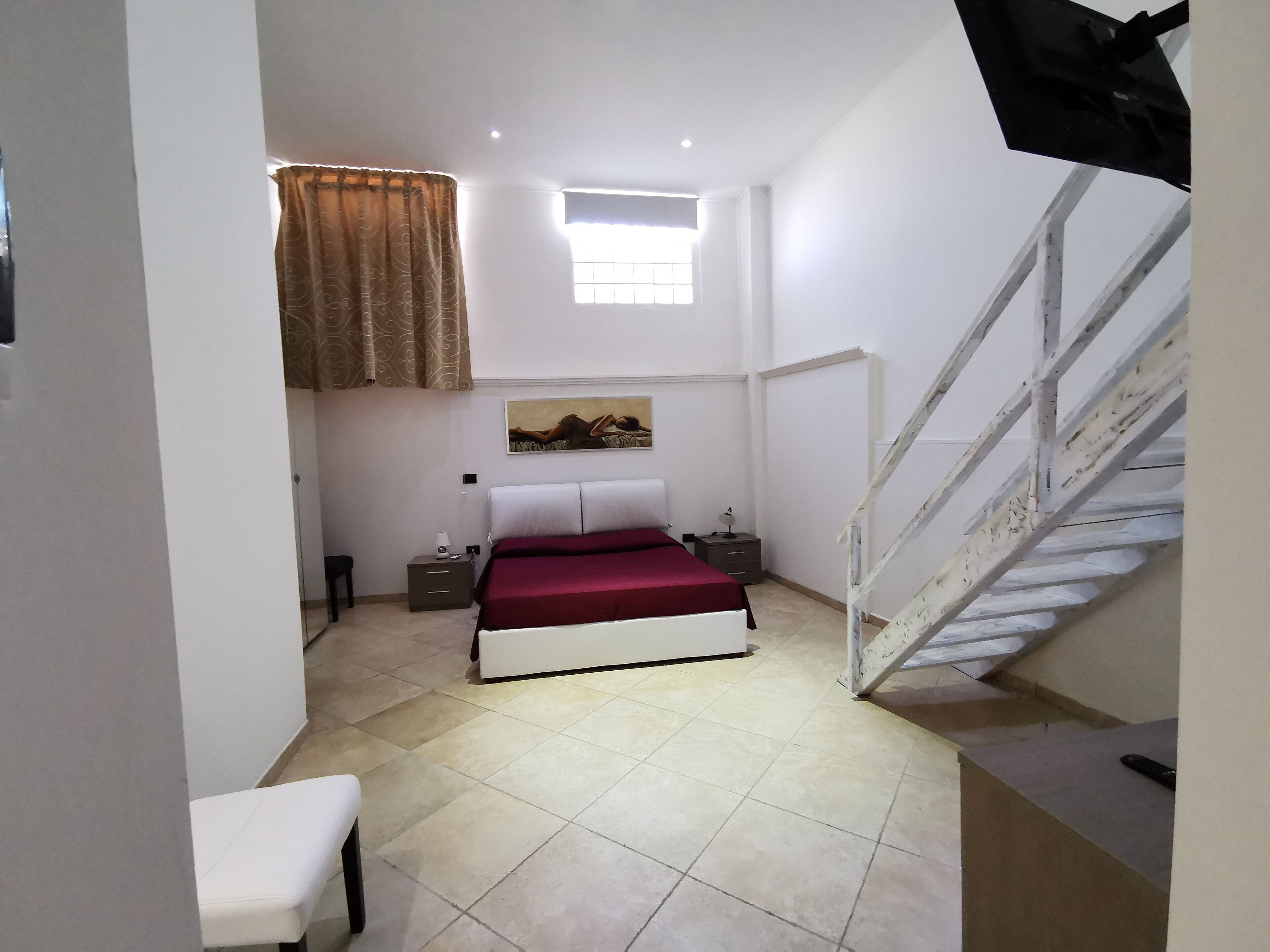A bedroom with a bed, a nightstand, a chair, a window with curtains, and a staircase.