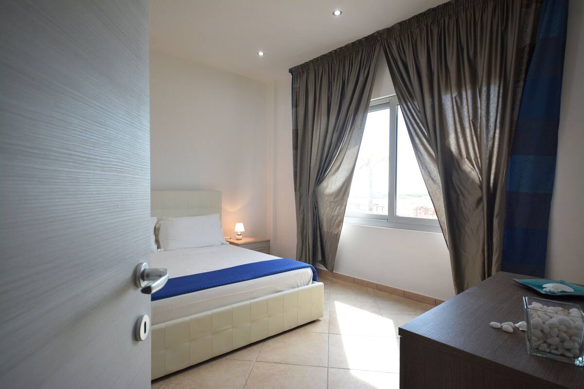 A hotel room with a bed, a nightstand, a desk with a lamp, and a window with curtains.