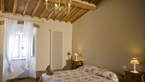 A bedroom with a wooden ceiling, a bed with floral bedding, two bedside tables with lamps, a radiator, and a window with curtains. - VILLA SOURROUNDED WITH VINEYARDS - BORGO CASANOVA (Pontassieve)