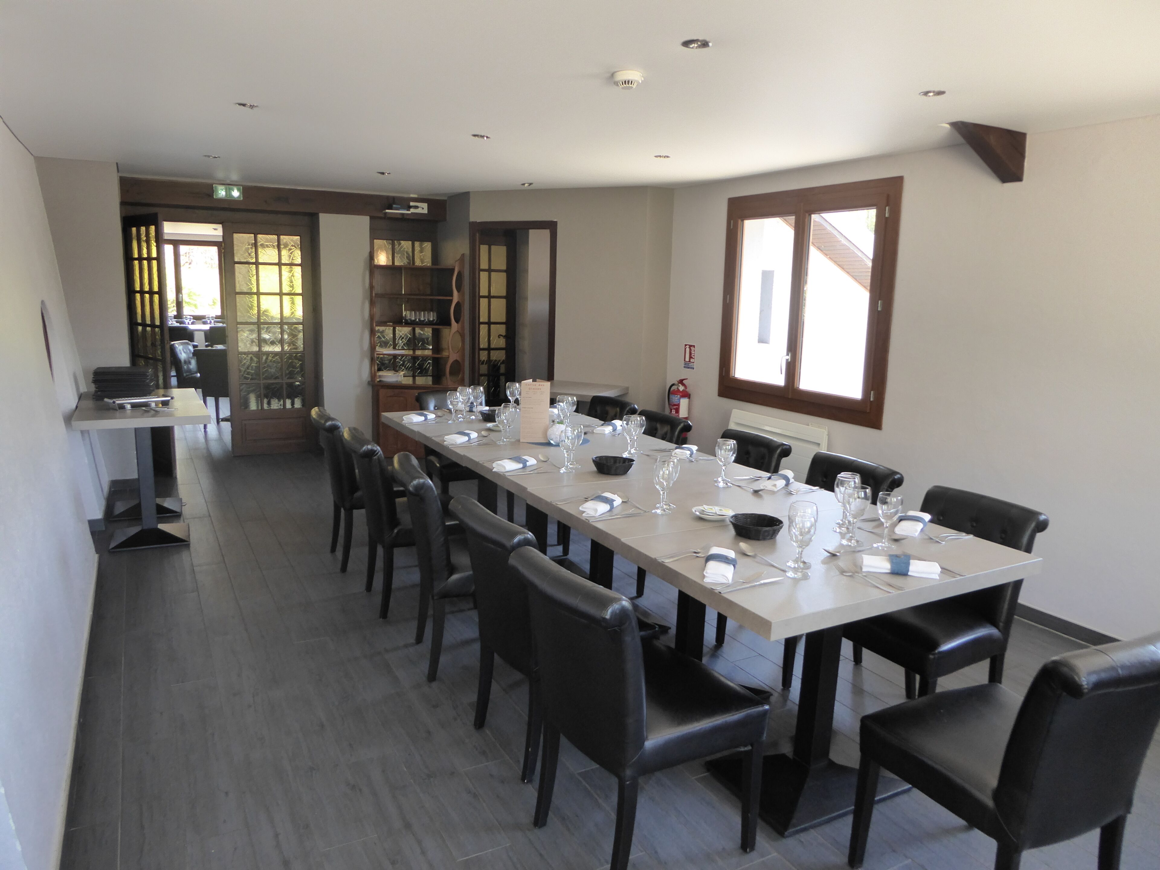 A conference room with a long table set for a meeting, surrounded by chairs, with a bookshelf and a fire extinguisher in the background.