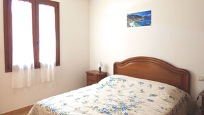 Un dormitorio con cama de madera, una mesita de noche con lámpara, una ventana con cortinas y un cuadro pequeño en la pared.