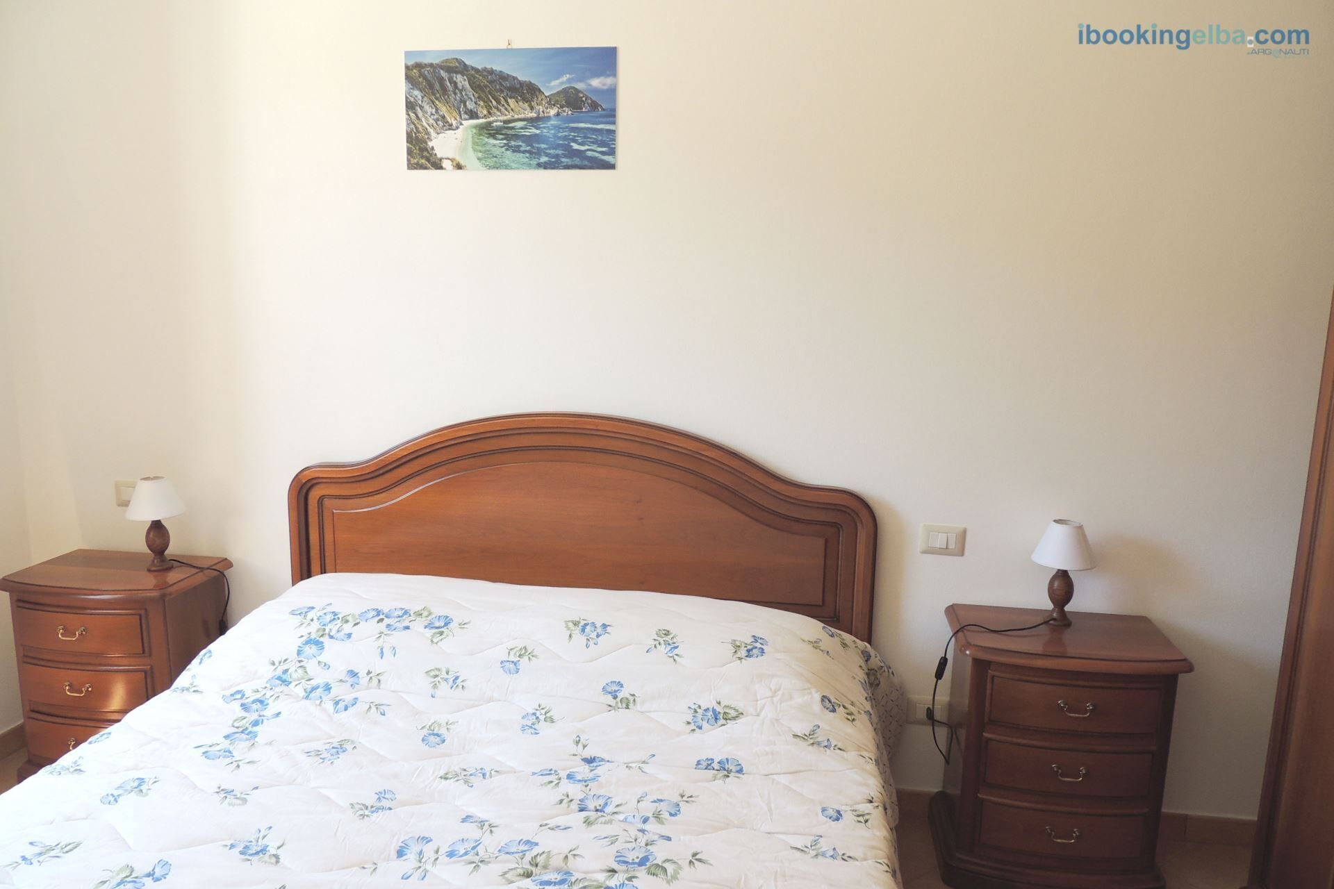 A bedroom with a wooden bed, a floral bedspread, two bedside tables with lamps, and a small picture on the wall.