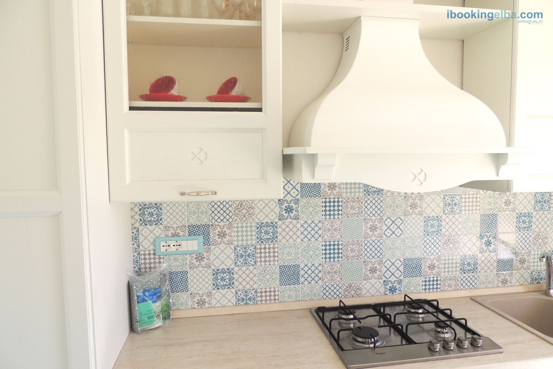 A kitchen with a gas stove, a tiled backsplash, and a white cabinet with a red hat on the shelf.