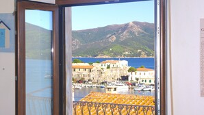 A view from a balcony with a metal railing, overlooking a marina with boats, a coastal town, and a mountain in the background. - Cosmopolis (Portoferraio)