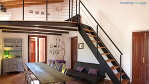 A spacious living area with a wooden dining table, clear chairs, a black sofa with cushions, a staircase with wooden steps, and a wall-mounted shelf with decorative items. - Cottage Vittoria (Portoferraio)