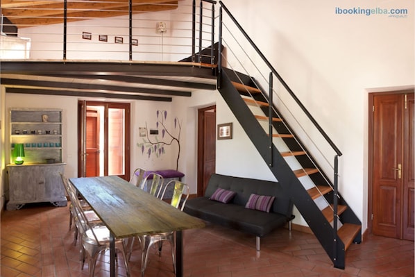 A spacious living area with a wooden dining table, clear chairs, a black sofa with cushions, a staircase with wooden steps, and a wall-mounted shelf with decorative items.