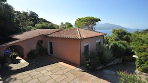 A single-story house with a tiled roof, a paved driveway, and a view of the sea and mountains in the background.