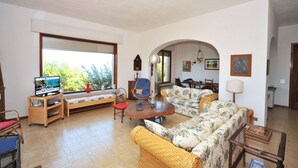 A living room with a sofa, armchairs, a small table, a TV, and a window with blinds.