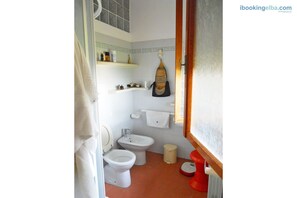 A bathroom with a toilet, bidet, and a red bidet seat. There are shelves with toiletries and a towel hanging on the wall. - Villa Enfola (Portoferraio)