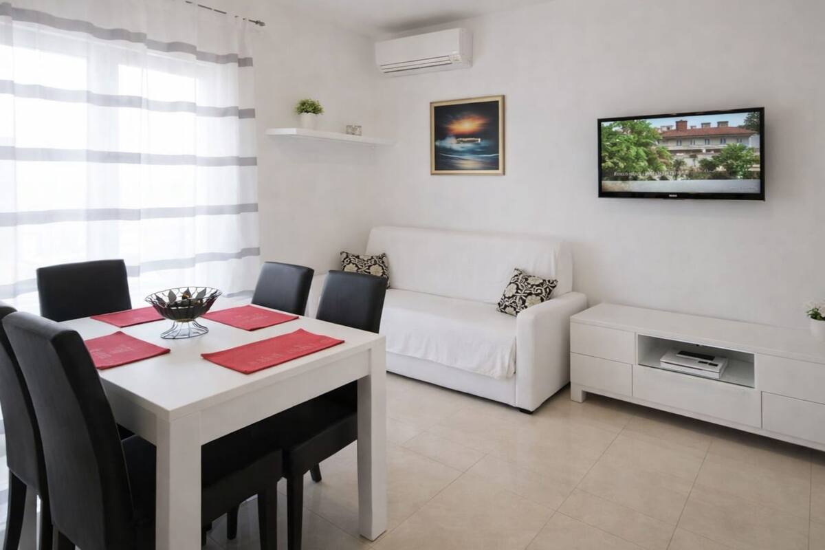 A modern living room with a white sofa, a dining table with chairs, a TV mounted on the wall, and a small shelf with a plant.
