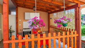 Une pergola en bois avec une table et des chaises en dessous, un pot de fleurs suspendu et, au loin, une maison.