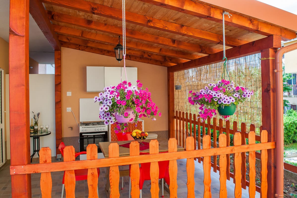 A wooden pergola with a table and chairs underneath, a hanging flower pot, and a view of a house in the background.