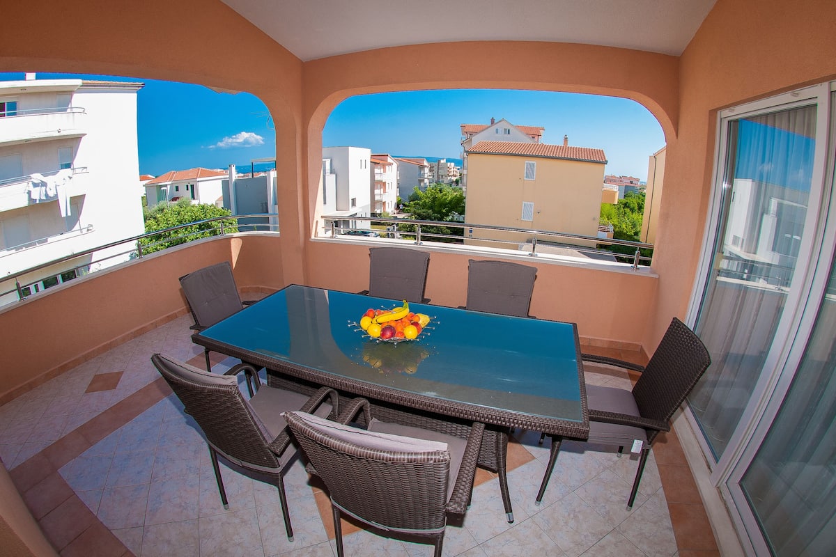 A balcony with a dining table and chairs, a fruit bowl, and a view of buildings and trees.