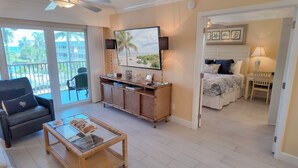 A spacious living area with a balcony, a dining area with a glass-top table, and a bedroom with a bed and bedside table.