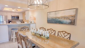 A dining area with a long table, chairs, and a chandelier. There is a kitchen in the background with modern appliances.