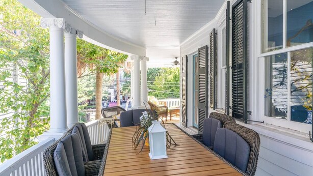 A covered porch with a wooden table and chairs, white columns, and shutters. - Historic Charm | The Bee & Blossom (Charleston)