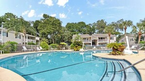 Ein Swimmingpool, umgeben von Liegestühlen und Palmen, mit einem Gebäudekomplex im Hintergrund.
