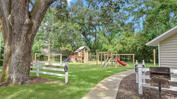 A playground with a slide and climbing structures, surrounded by trees and a white fence.