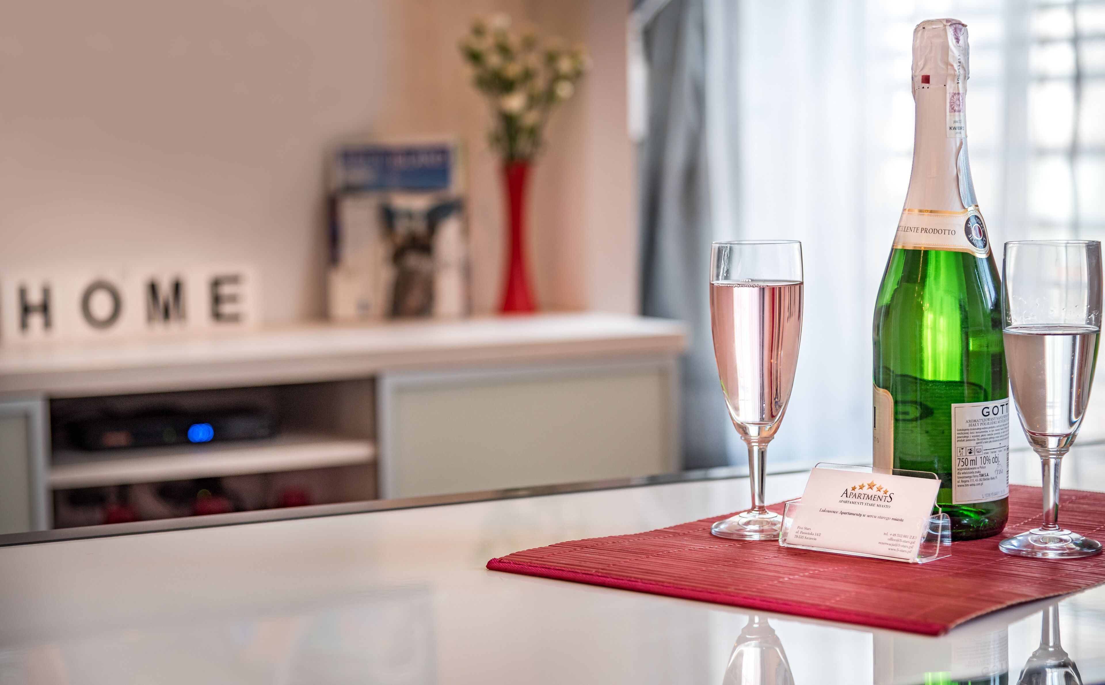 Habitación de hotel con un minibar, un mantel individual rojo con una botella de vino y copas, y un cartel con la palabra 'HOME'.