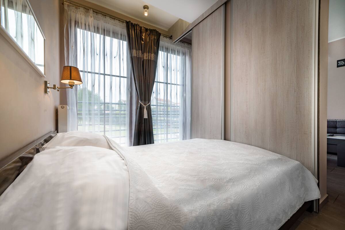 Une chambre d’hôtel avec un grand lit, une fenêtre avec des rideaux légers, une lampe fixée au mur et une vue sur l’extérieur.