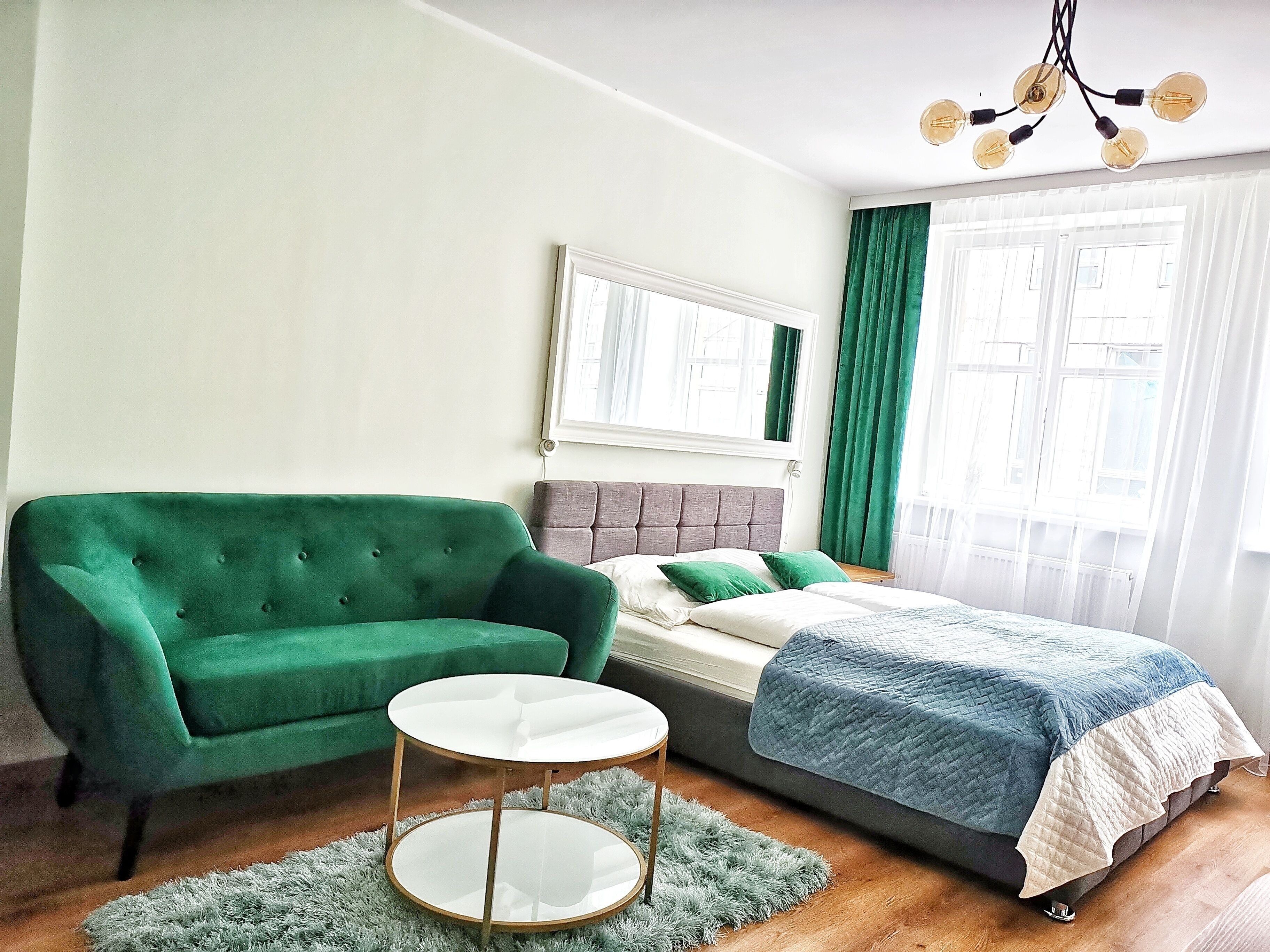 A bedroom with a green sofa, a round white table, a bed with a blue quilt, and a large window with curtains.