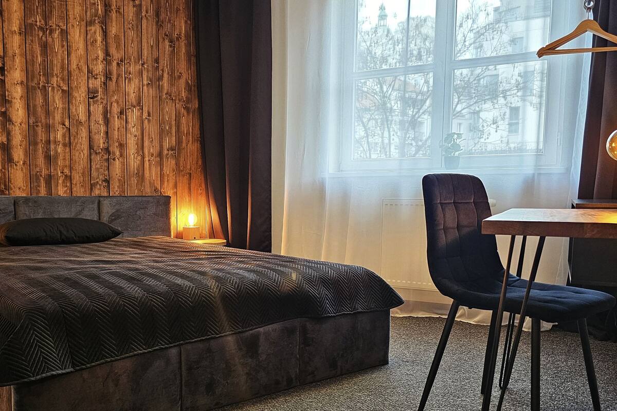 Une chambre à coucher avec une tête de lit en bois, un lit recouvert d’une couvre-lit à motifs, une chaise, un bureau et une fenêtre donnant sur des arbres.