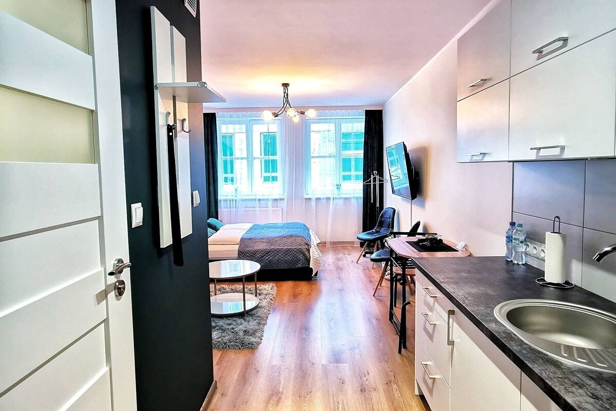 Une cuisine moderne avec des armoires blanches, un comptoir noir et un évier en acier inoxydable. On y trouve un coin repas avec une table et des chaises, et un lit à l’arrière-plan.