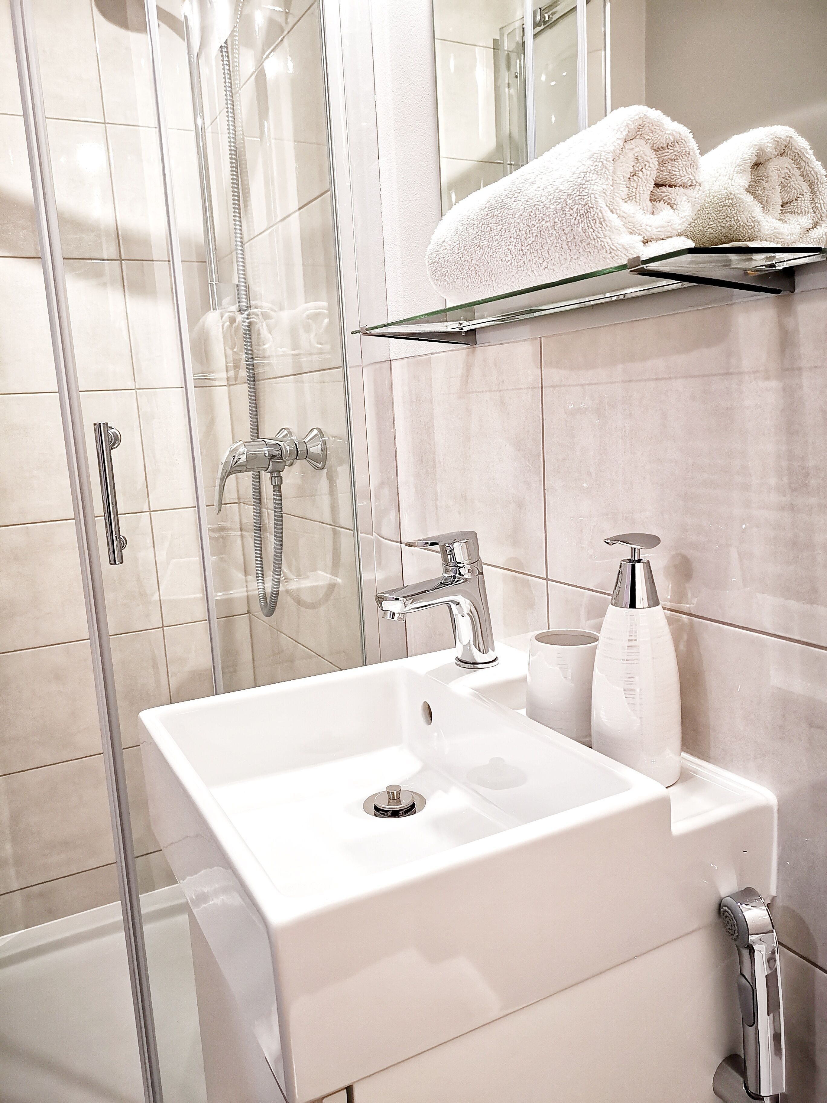 Ein modernes Badezimmer mit Duschkabine aus Glas, einem weißen Waschbecken mit Chromarmatur und einem Ablagebrett mit gerollten Handtüchern.