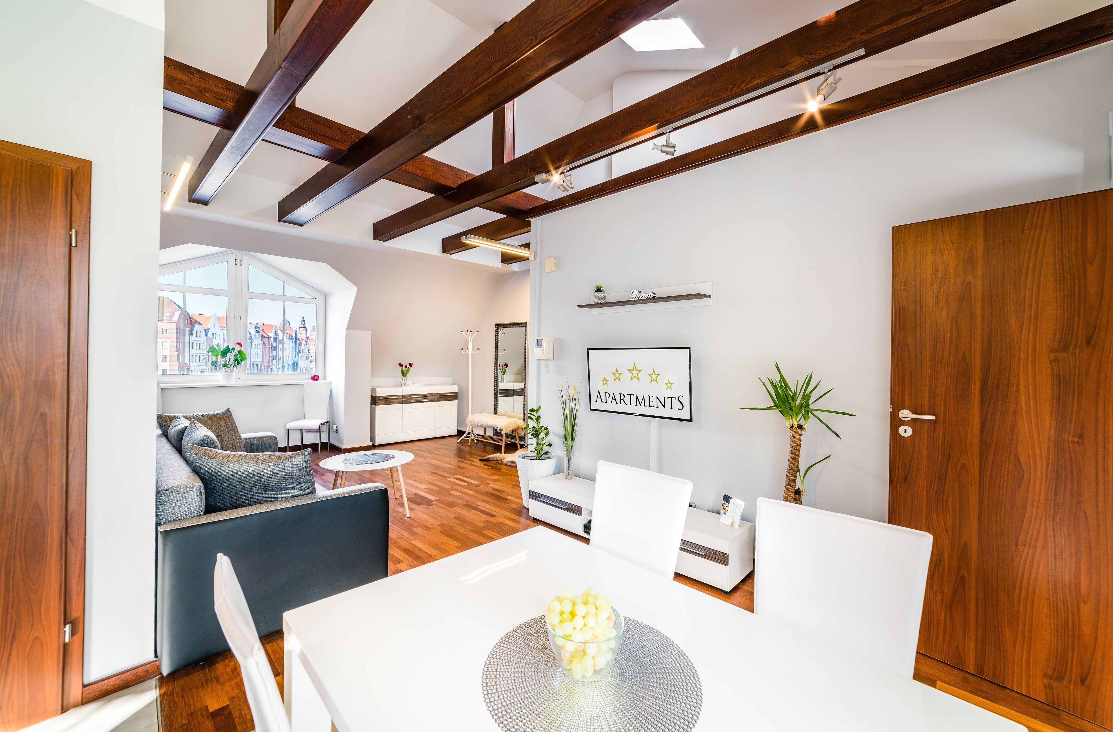 Ein modernes Wohnzimmer mit Essbereich, Holzbalken an der Decke und einem großen Fenster. Ein Schild zeigt die Aufschrift "Apartments".