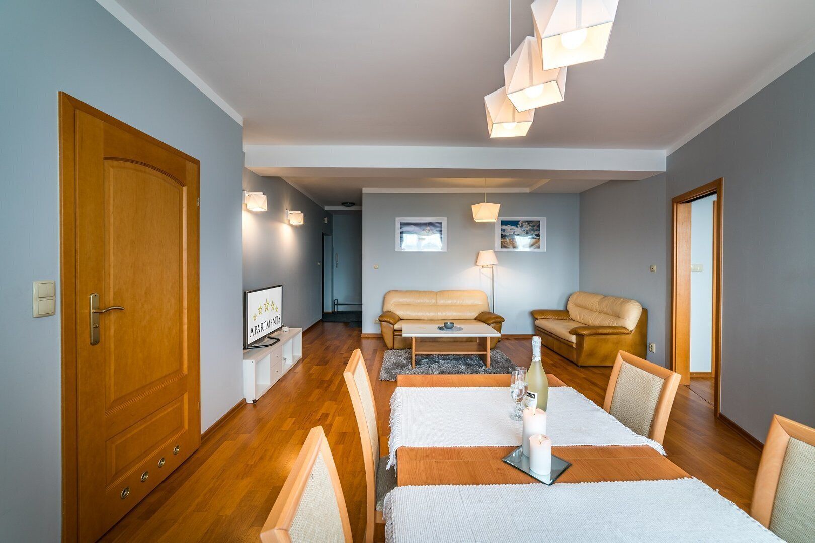 Een moderne woonkamer met houten vloer, een bank, een salontafel en een eettafel met stoelen. Links een deur en rechts een raam.