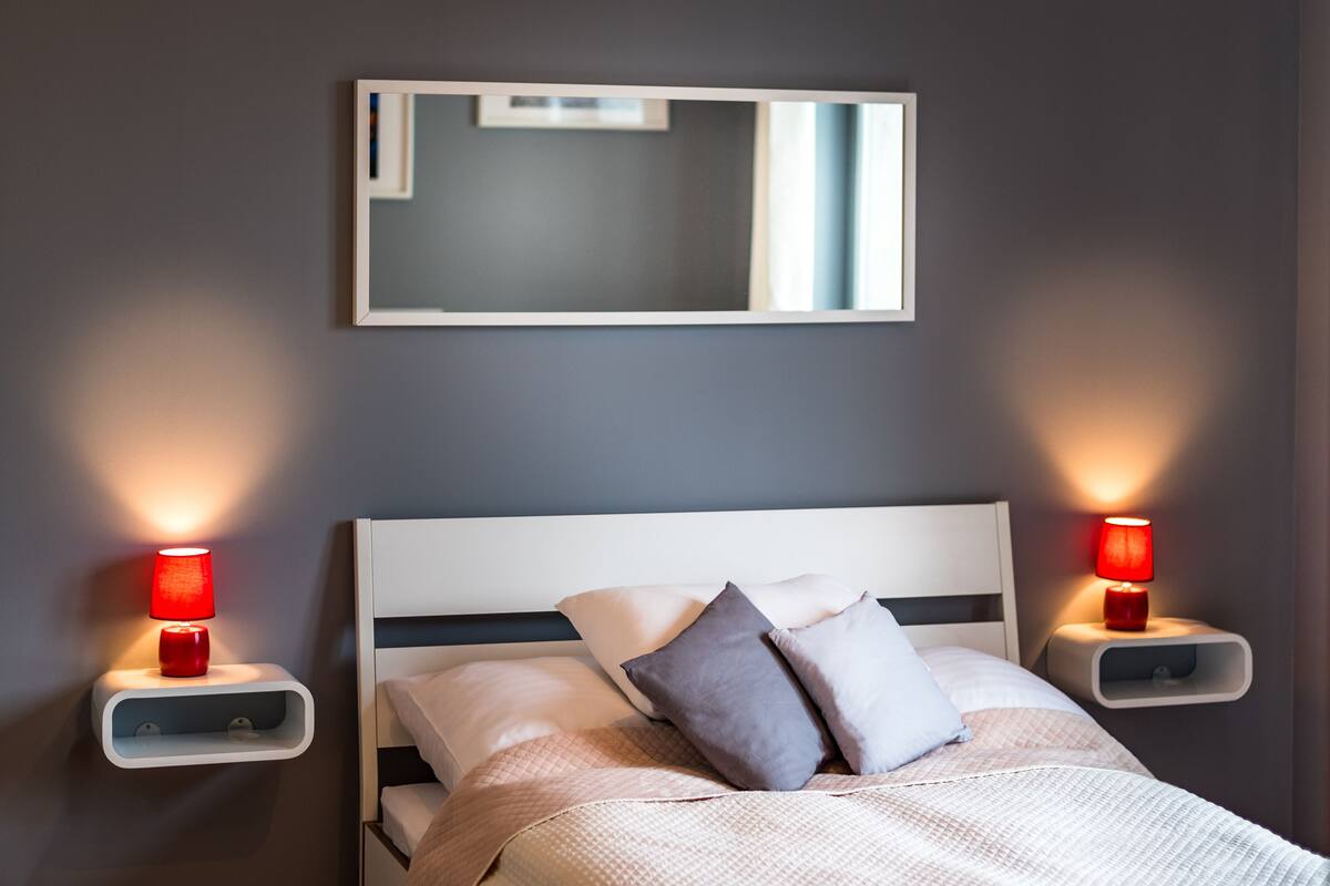 Une chambre moderne avec un lit blanc, deux tables de chevet avec des lampes rouges, un grand miroir au-dessus du lit et un mur de couleur sombre.