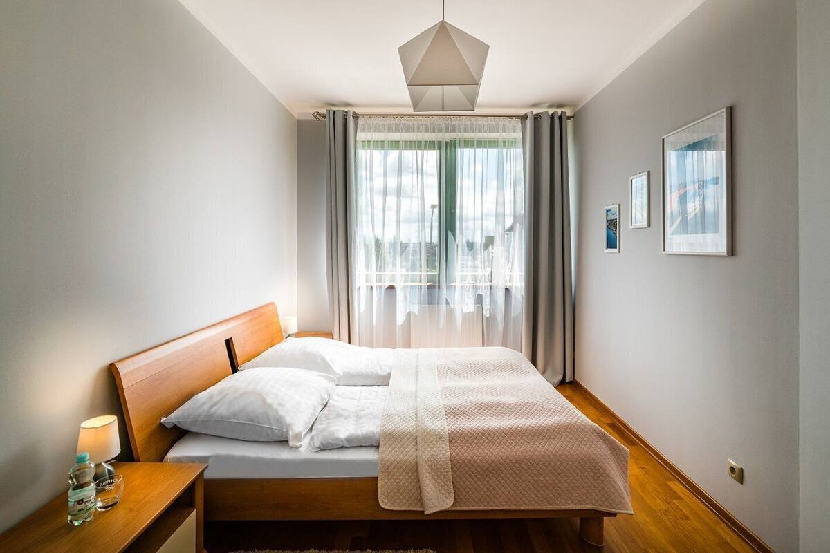 Une chambre avec un lit en bois, une table de chevet avec une lampe et une bouteille d’eau, un luminaire suspendu et une vue sur l’extérieur à travers des rideaux légers.