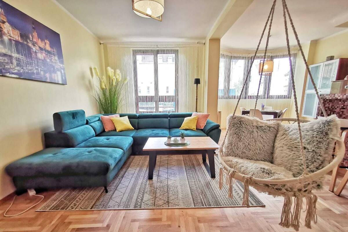 Un salon comprenant un canapé d’angle, un fauteuil suspendu, une table basse et une grande fenêtre avec des rideaux.