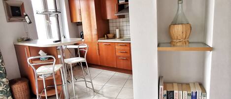 A modern kitchen with wooden cabinets, a dining area with metal chairs, and a bookshelf with books and decorative items.