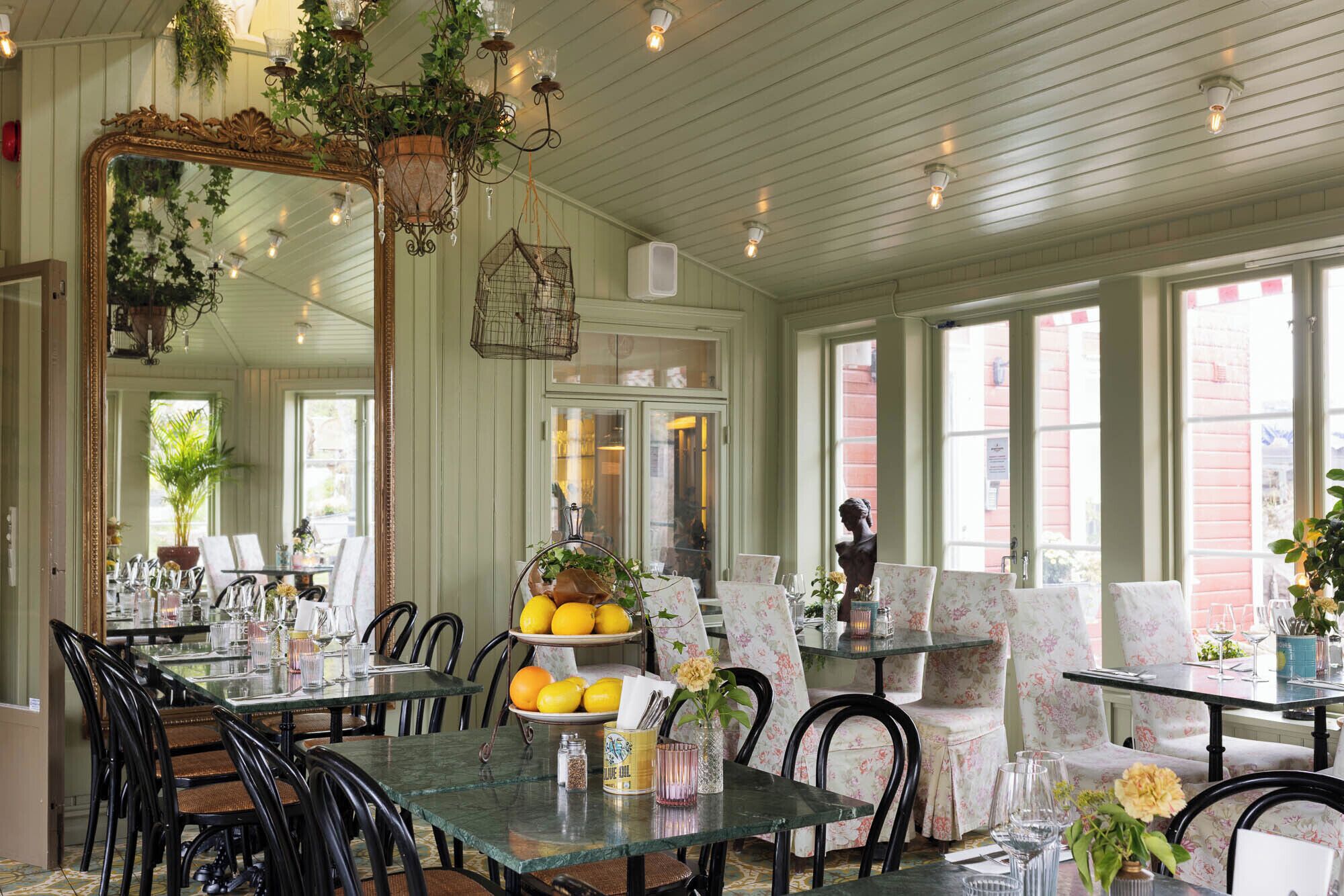 A dining area with glass tables, black chairs, and a large mirror. There are potted plants and a hanging light fixture.