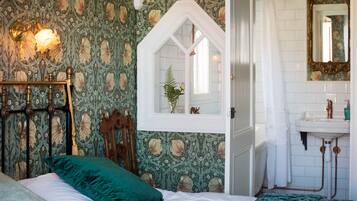 Un dormitorio con cama, ventana con vista a una planta, baño con lavamanos y espejo, y pared con papel tapiz floral.