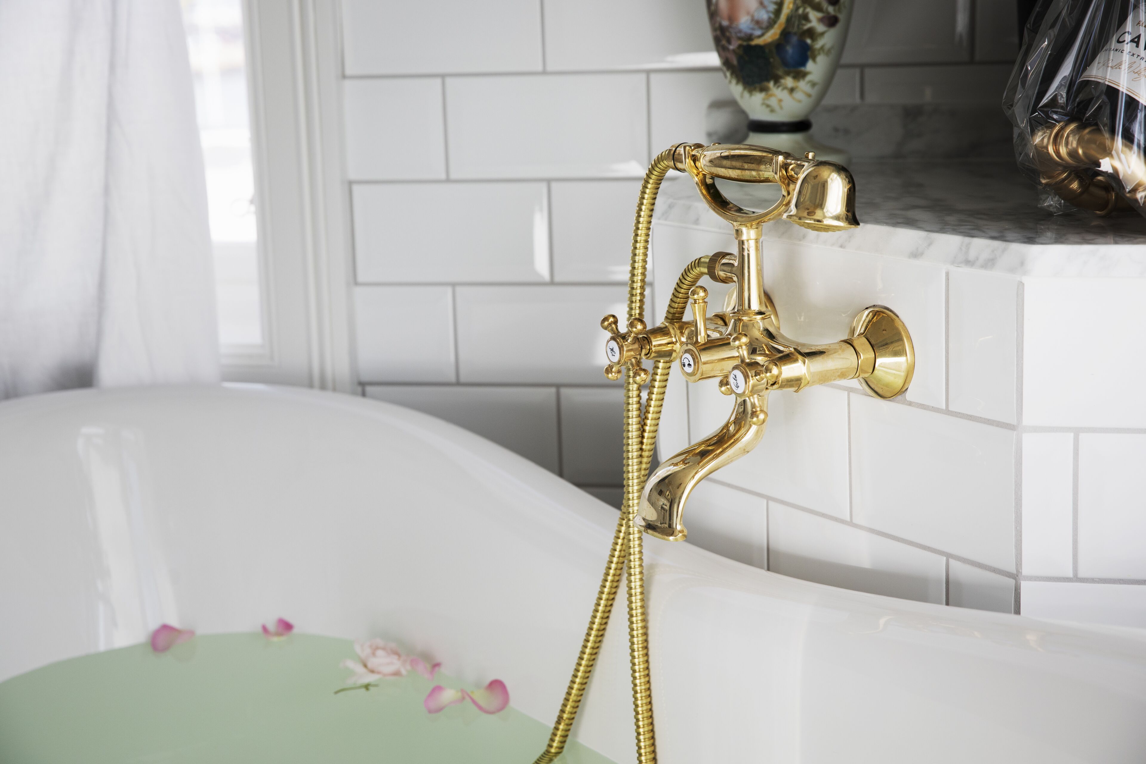 A bathtub with a gold faucet and shower head, white tiled walls, and a decorative vase in the background.