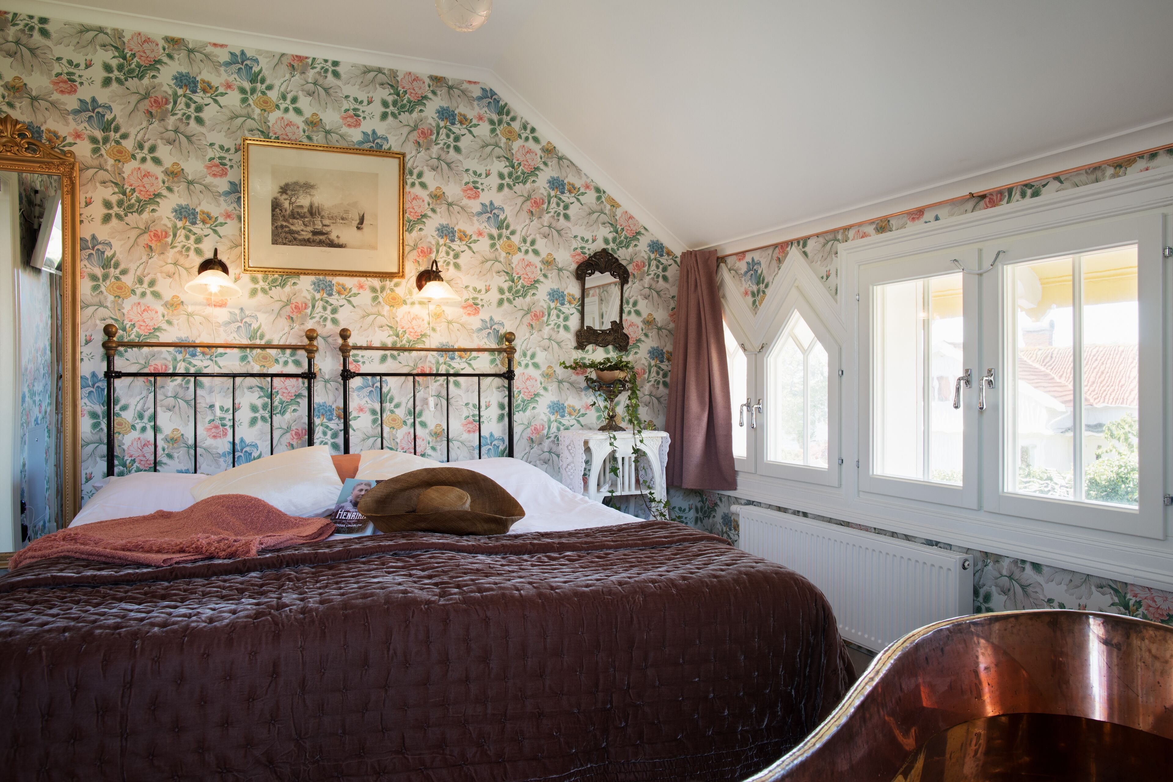 A bedroom with a floral wallpaper, a bed with a brown quilt, a wooden headboard, a nightstand, a mirror, and a window with curtains.