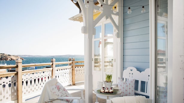 A balcony with a wooden deck, white chairs, a small table with a plant and a plate of food, and a view of the sea. - Strandflickornas Annex Gula Villan (Lysekil)