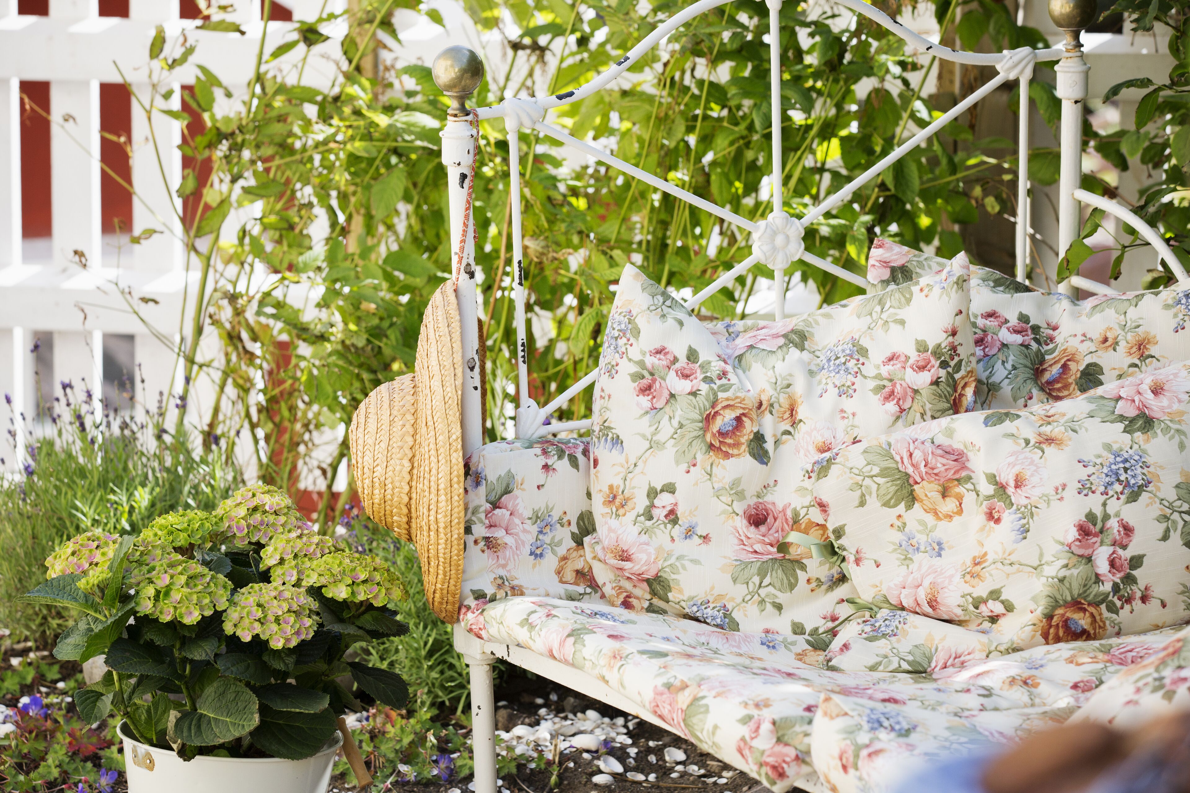 Een witte metalen schommelstoel met bloemenstoffering, een geweven mand en een potplant met groene bladeren en paarse bloemen.