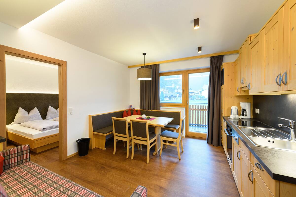 Quarto compacto com cama, mesa de jantar com cadeiras, pequena cozinha e vista pela janela.