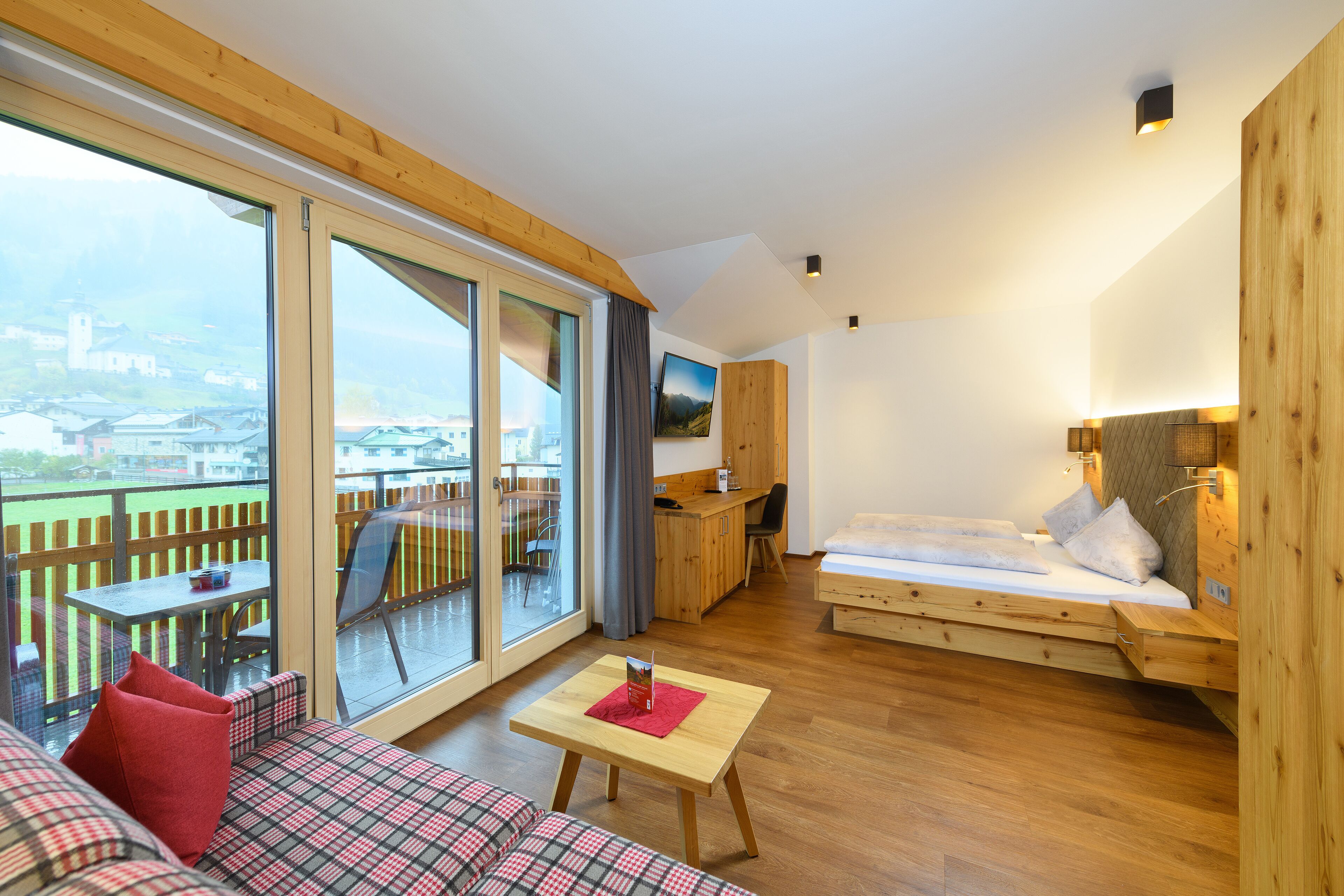 A modern room with a large sliding glass door leading to an outdoor area, a wooden bed, a small table, and a sofa with plaid upholstery.