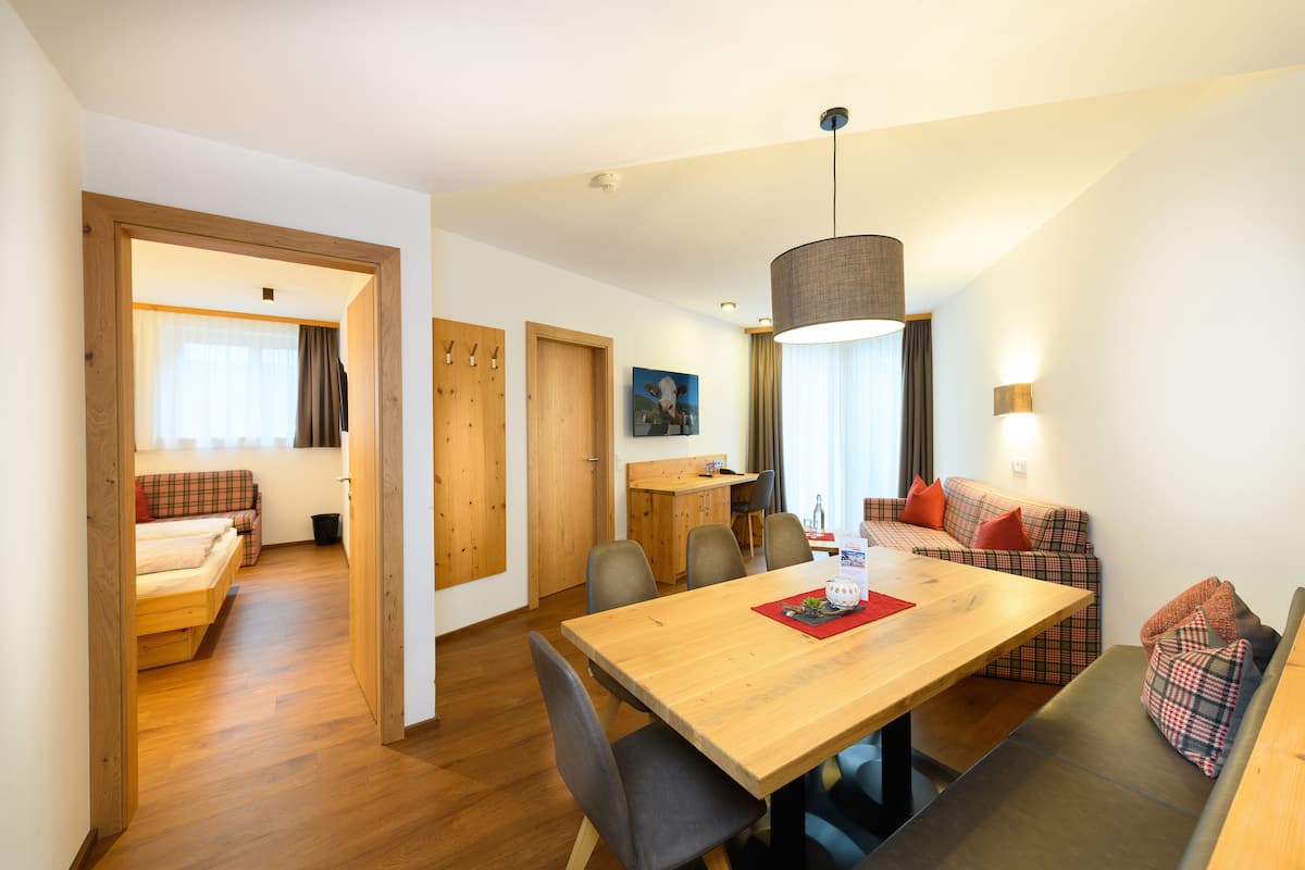 Quarto moderno e bem iluminado, com uma mesa de jantar de madeira, um sofá e um banquinho. Há uma porta que dá para outro quarto com uma cama e uma janela com cortinas.