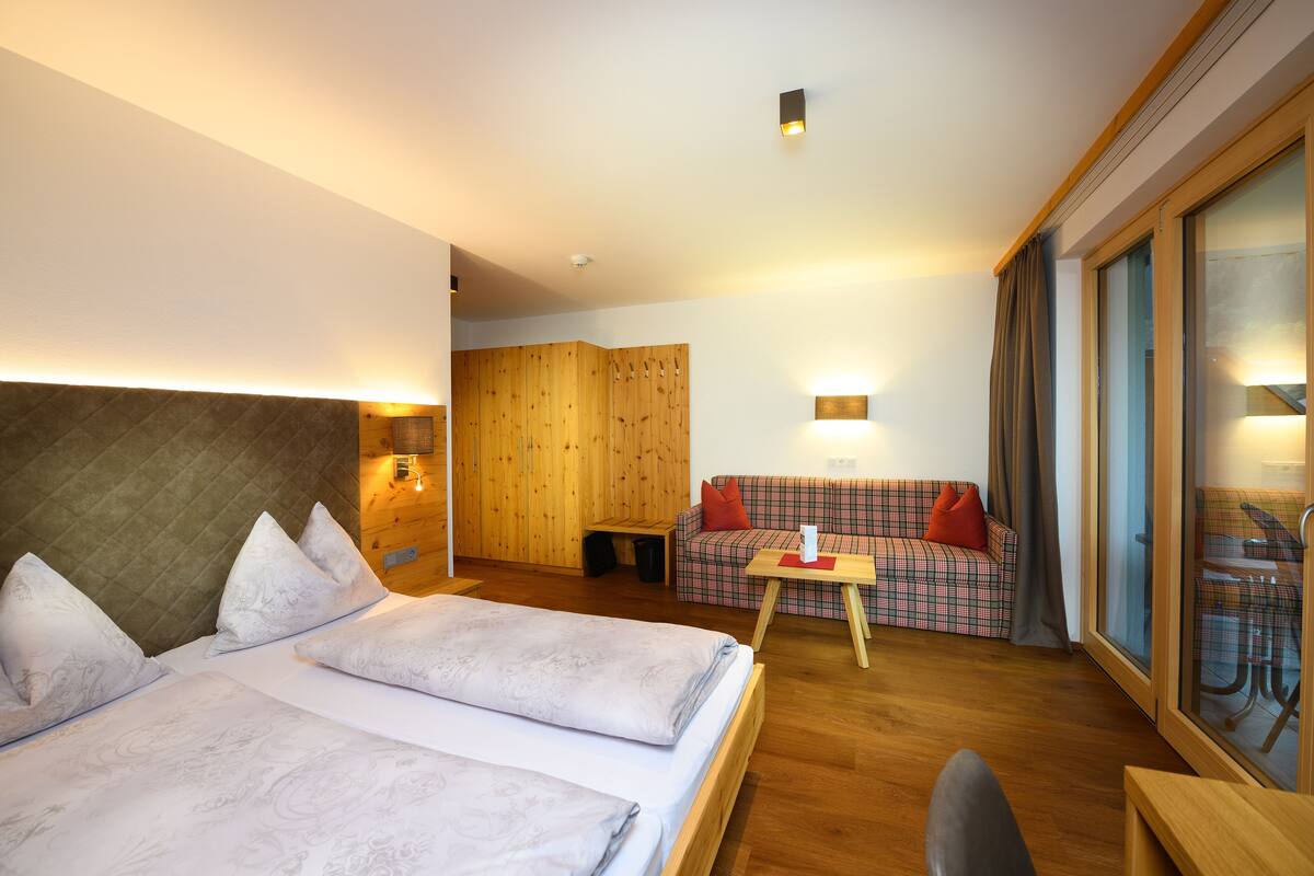 Quarto de hotel moderno com duas camas, um sofá, uma mesinha e uma porta de vidro deslizante para uma varanda.