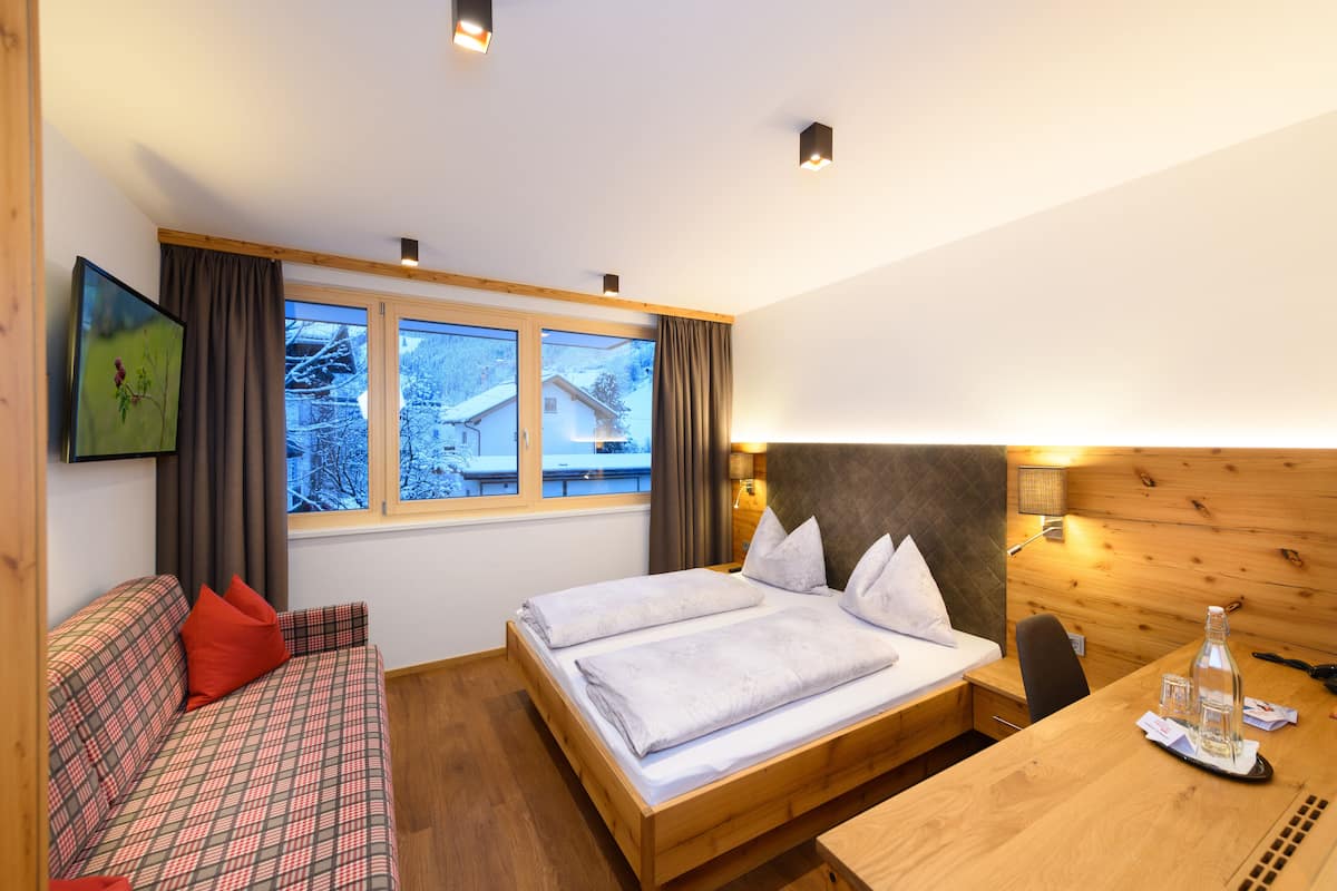 Quarto de hotel moderno com cama de madeira, sofá com almofadas vermelhas, escrivaninha de madeira e uma grande janela com vista para uma paisagem nevada.