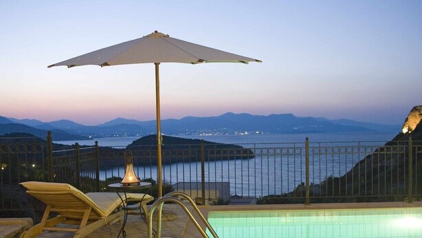 A poolside area with a view of the sea and mountains, a lounge chair, an umbrella, and a table with a lamp. - Villa Helios with private pool, beautiful sea view and sunny terrace (Agios Nikolaos Lassithi)