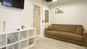 Un salón moderno con un televisor de pantalla plana, un sofá marrón, una estantería blanca con cristalería y una pared de ladrillo decorativa.