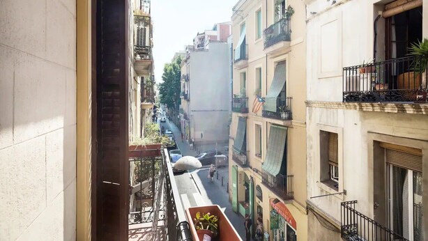 A narrow street between buildings with balconies, some with plants and flags. - Stay U-nique Apartment Travessera (Barcelona)