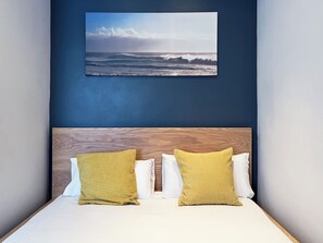 Ein Schlafzimmer mit einem Holz-Kopfteil, zwei gelb gemusterten Kissen und einem weißen Kissen. Über dem Bett hängt ein Gemälde, das eine Meereslandschaft zeigt.