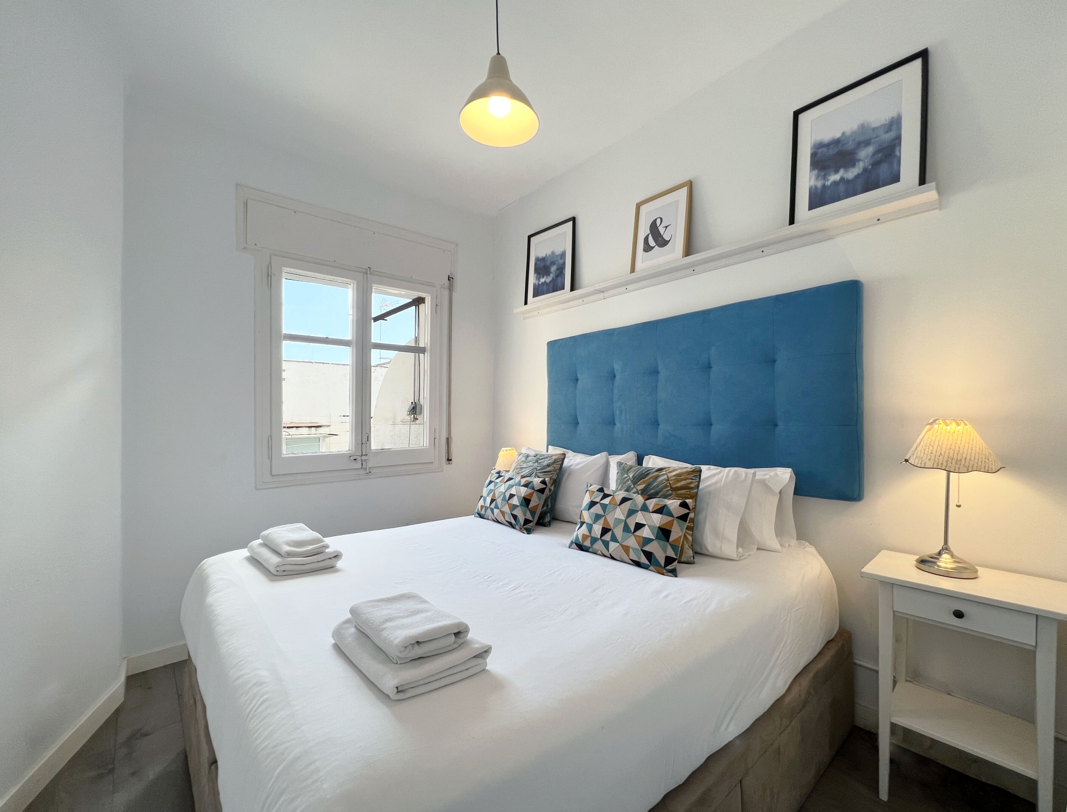 A neatly made bed with white linens and folded towels, a blue upholstered headboard, and a bedside lamp on a white nightstand.